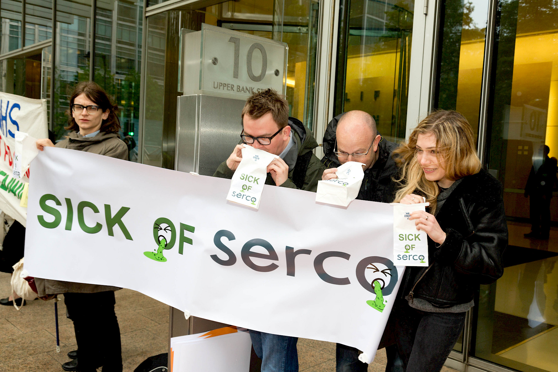 People holding a Sick of SERCO banner in front of an office building.