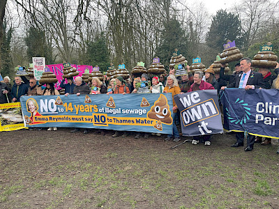 A crowd of protestors. In the foreground, protestors hold banners saying 'No to 14 years of illegal sewage: Emma Reynolds must say NO to Thames Water'; 'We Own It' and 'the Climate Party'. In the  background, protestors hold up 14 giant inflatable poos, each with a crown and a year on it