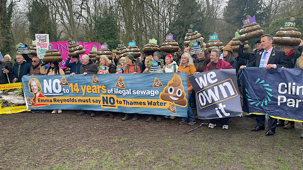 A crowd of protestors. In the foreground, protestors hold banners saying 'No to 14 years of illegal sewage: Emma Reynolds must say NO to Thames Water'; 'We Own It' and 'the Climate Party'. In the  background, protestors hold up 14 giant inflatable poos, each with a crown and a year on it