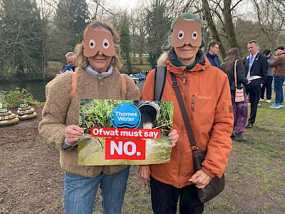 Two protestors with masks in the shape of poo emojis. They are holding a sign which says 'Ofwat must say NO'. In the background of the sign, a pipe leaks sewage into a  river.