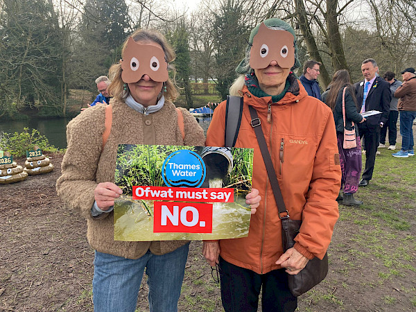 Two protestors with masks in the shape of poo emojis. They are holding a sign which says 'Ofwat must say NO'. In the background of the sign, a pipe leaks sewage into a  river.