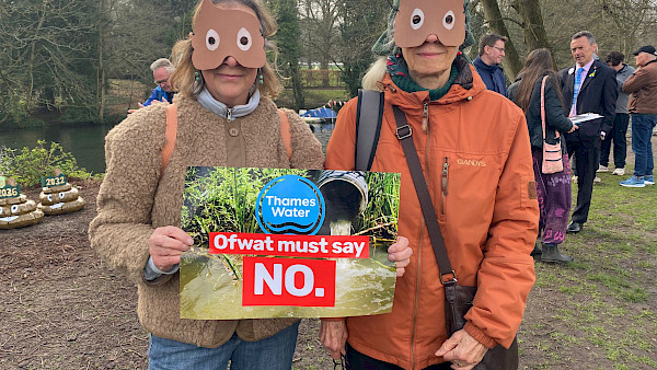 Two protestors with masks in the shape of poo emojis. They are holding a sign which says 'Ofwat must say NO'. In the background of the sign, a pipe leaks sewage into a  river.