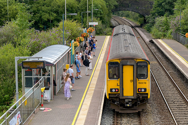 Photograph, shot from above, of passengers waiting to board a train as it pulls into a small railway station in the countryside.