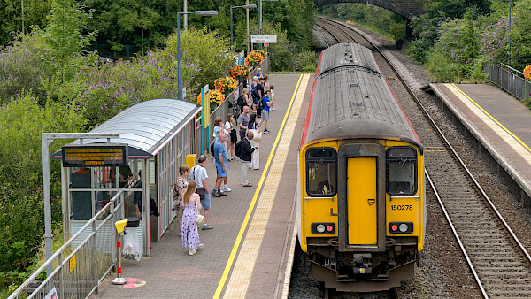 Photograph, shot from above, of passengers waiting to board a train as it pulls into a small railway station in the countryside.