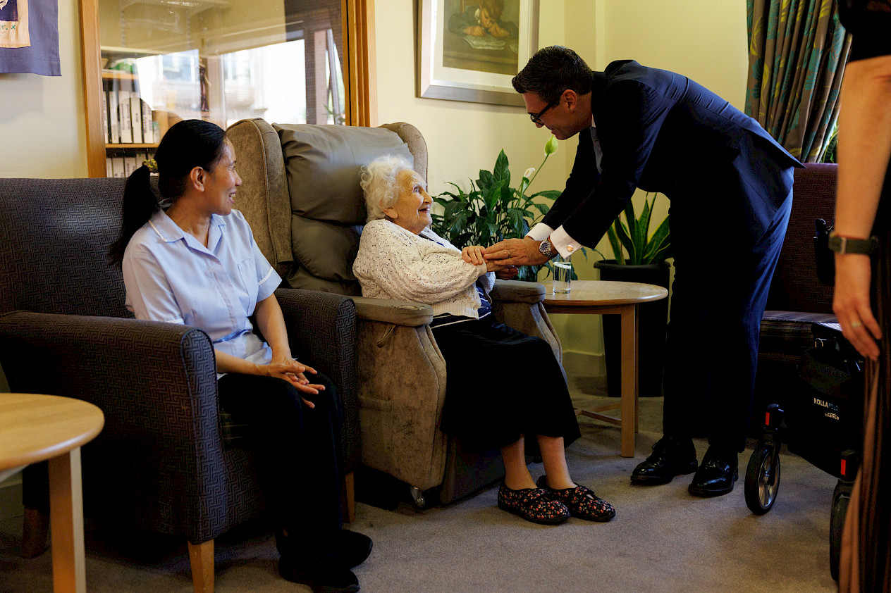 Photo of Burnham in Jewish Care. He's bending down to hold an old woman's hands, she is smiling up at him. A care worker is sat smiling in the next seat.