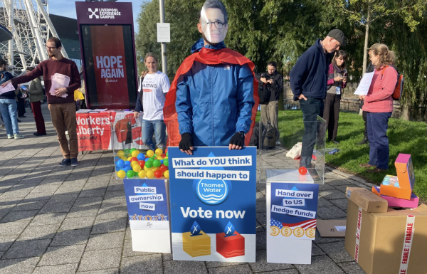 Image of a vote outside Labour Party conference. There are two clear perspex boxes, one is full of coloured balls and says 'public ownership now'. The other contains only one ball and says 'hand over to US hedge funds'. In the middle, a man stands with an Andy Burnham mask, a cape and a sign which reads: What do YOU think should happen to Thames Water - vote now'