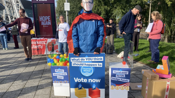 Image of a vote outside Labour Party conference. There are two clear perspex boxes, one is full of coloured balls and says 'public ownership now'. The other contains only one ball and says 'hand over to US hedge funds'. In the middle, a man stands with an Andy Burnham mask, a cape and a sign which reads: What do YOU think should happen to Thames Water - vote now'