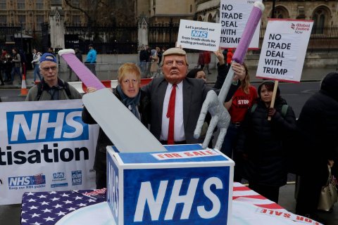 An image of a protester dressed as Donald Trump, carving up a box labelled 'NHS' outside Parliament, London