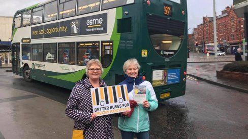 Two women hold a placard next to a bus saying 'Better buses for greater Manchester'