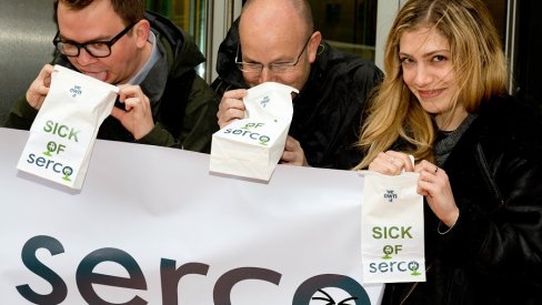 Image of 3 people holding a banner saying 'Sick of Serco', and miming being sick into paper bags